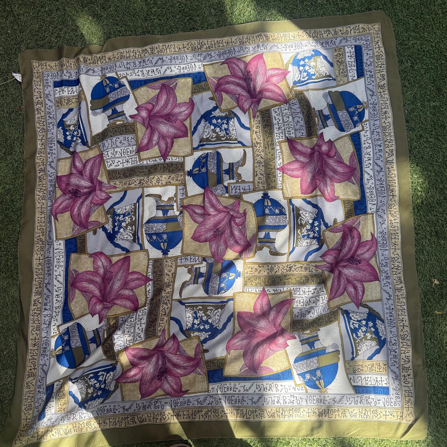 A square silk scarf with a traditional Persian-style pattern featuring pink lotus flowers, blue and white architectural motifs, and gold borders, laid out on green grass with a person's shoes visible at the bottom.