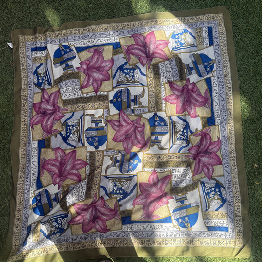 A square silk scarf with a traditional Persian-style pattern featuring pink lotus flowers, blue and white architectural motifs, and gold borders, laid out on green grass with a person's shoes visible at the bottom.